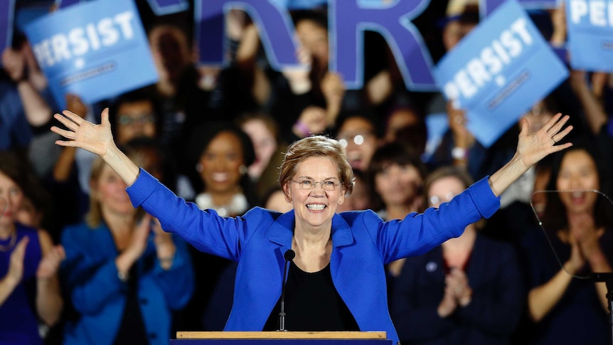 Elizabeth Warren stands at a podium in front of hundreds of supporters at a political rally, with her arms outreached