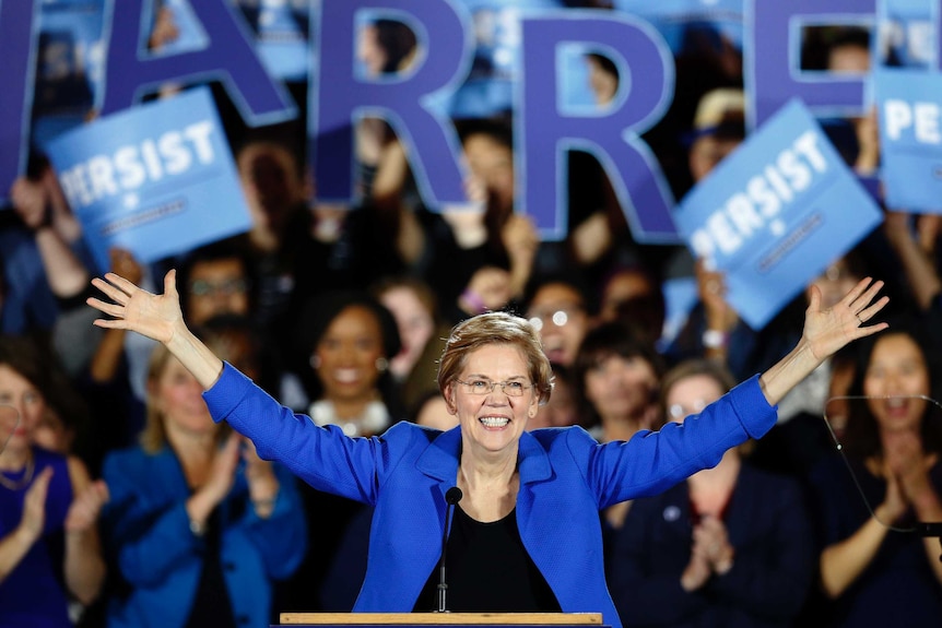 Elizabeth Warren stands at a podium in front of hundreds of supporters at a political rally, with her arms outreached