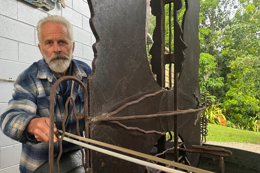 An older man with white hair and beard uses a musical bow on a large metal sculpture 