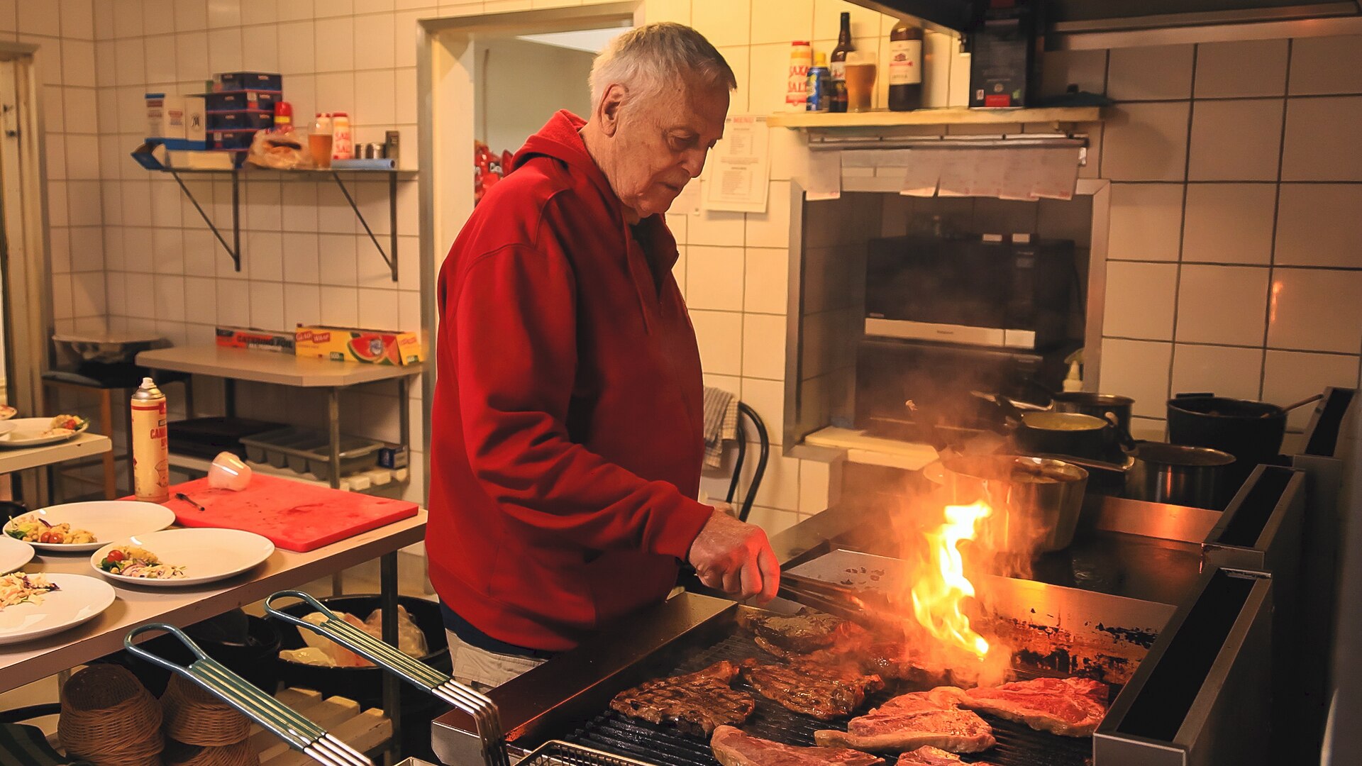 An old man cooks a steak in an industrial kitchen