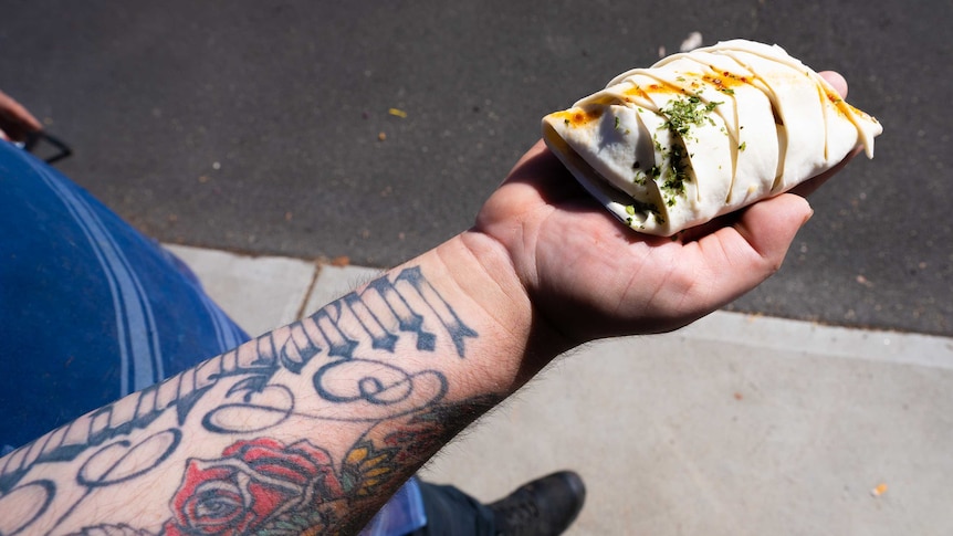 An arm lined with colourful tattoos holds out a pastry meat parcel into the sunlight.