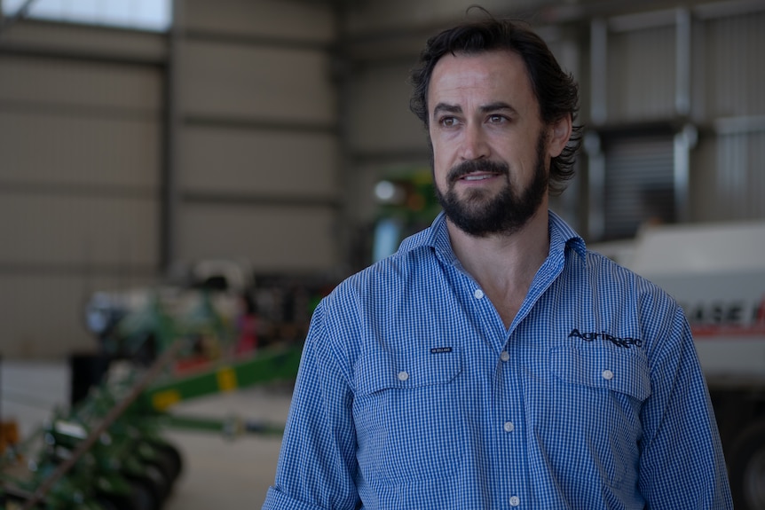 A man with dark hair and a blue shirt stands in an expansive but empty machinery dealer's shed.