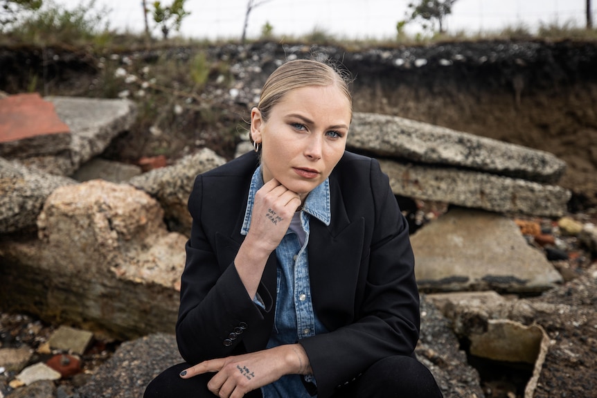 A blonde woman in a black blazer sits with her hand on her chin