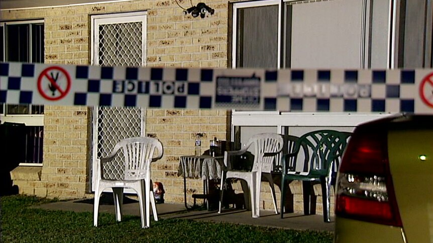 Police tape in front of a brick home. Plastic chairs, a door and windows are visible.