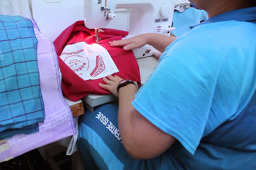 A woman sits at a sewing machine, she is wearing the Townsville Correctional Centre uniform and sewing a Boomerang Bag