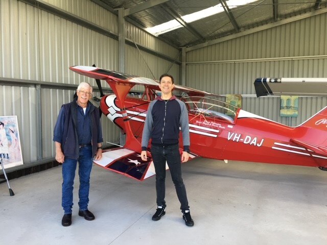An older man with white hair stands next to a younger tall boy in front of a red aerobatics plane in a hangar
