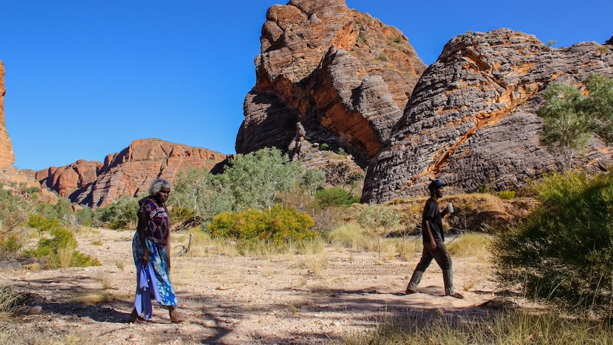 An older woman in a blue skirt, walking in front of a large rock in a national park