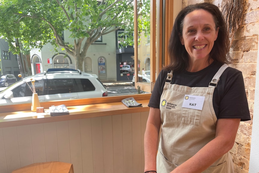 A woman wearing an apron smiles in front of an open window looking on to a leafy street