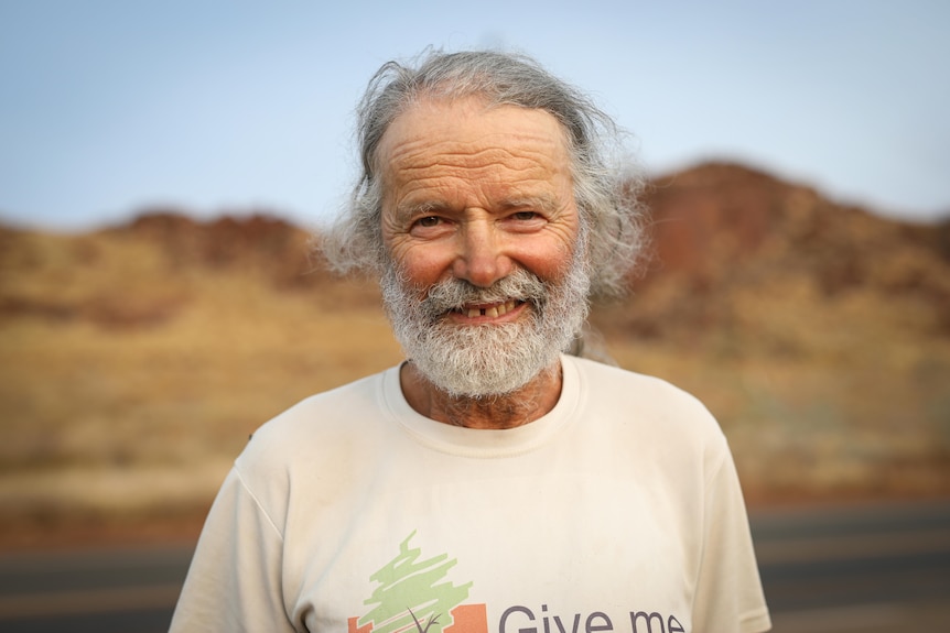 An older man with long grey hair smiles towards the camera and stands in front of red Pilbara hills