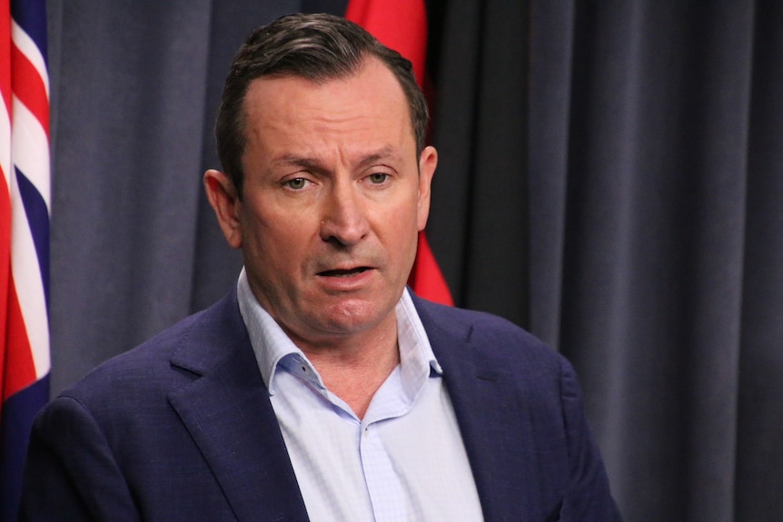 Mark McGowan standing in front of an Australian flag, wearing a navy blue suit and light blue shirt.