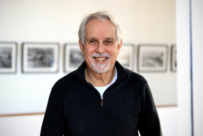 David Roach smiles wearing a black jumper with historical photos hanging behind him.