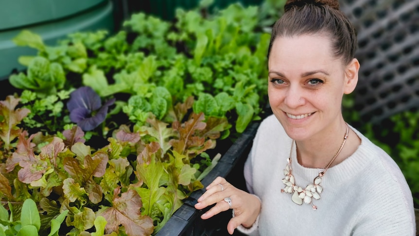 A woman looks at the camera while leaning beside a vegetable box of home grown lettuce