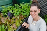 A woman looks at the camera while leaning beside a vegetable box of home grown lettuce