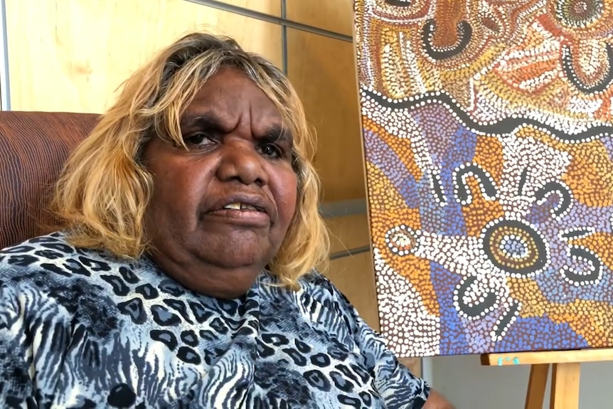 Deborah Walkabout, an Indigenous woman, sits in a chair in front of a painting.