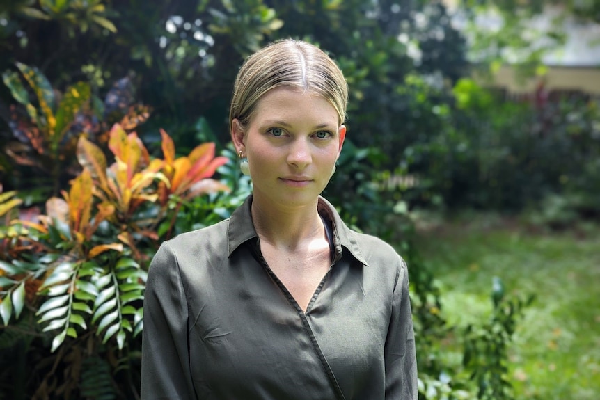 A woman with blonde hair wearing a green button up shirt in front of plants in a backyard