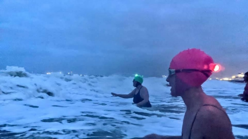 Three people wearing swimming caps in the whitewash of the ocean at dawn.