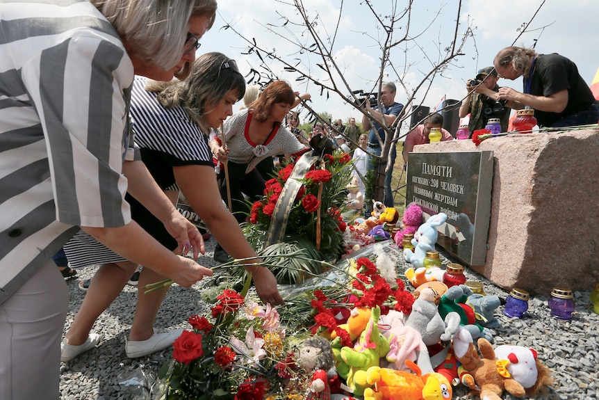 People bend down to place flowers at a gravestone where several stuffed toys and candles already sit