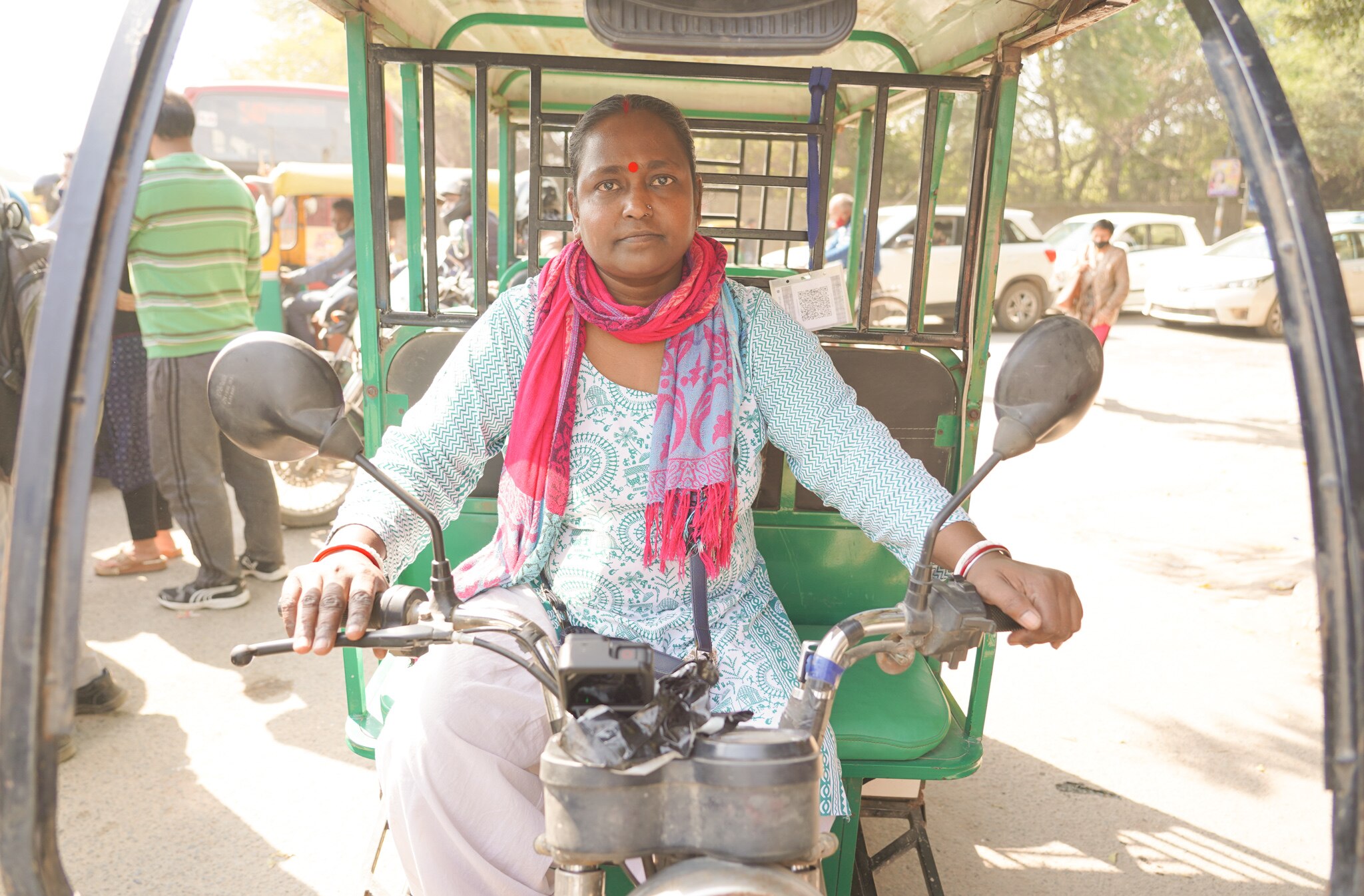 A woman wearing a pink scarf, blue top and khaki pants sits on a rickshaw