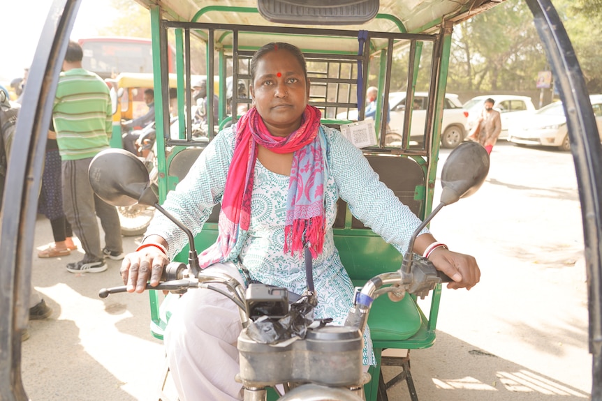 A woman wearing a pink scarf, blue top and khaki pants sits on a rickshaw