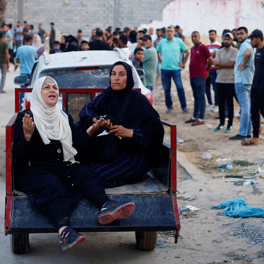 Two older Palestinian women, one crying out, sit on the back of a trailer as it traverses a crowded, dirty street.