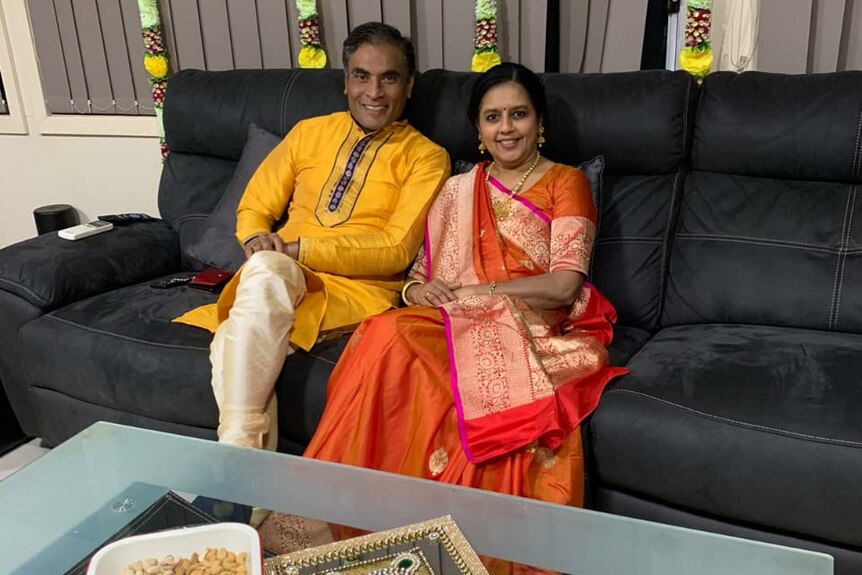 A man and woman sit on a black sofa, with trays of chocolate and Indian sweets on the table in front of them.