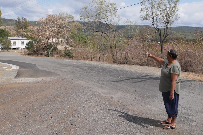 A woman stands on a bitumen road. She is wearing a green top and navy pants, and is pointing at land in the background.