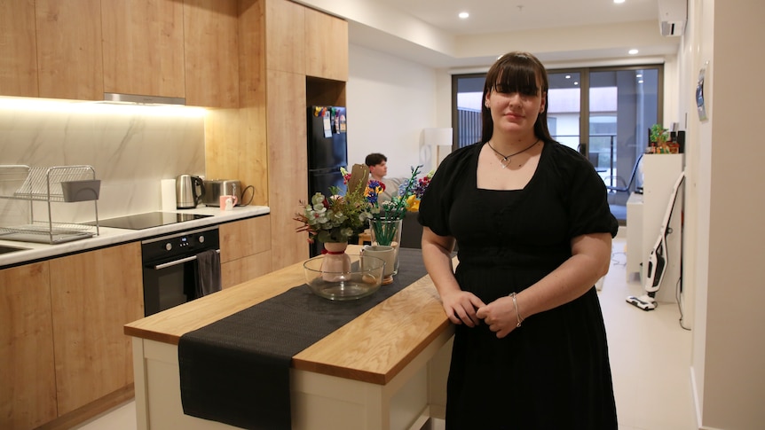 A young woman with dark hair and a black dress stands in a softly lit Melbourne apartment.