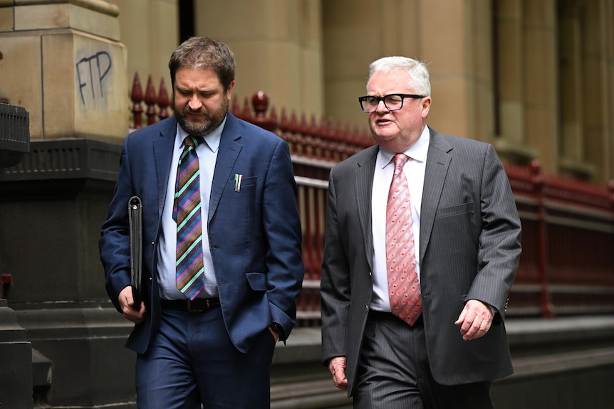 Two men wearing a blue and grey suit walk outside court buildings.