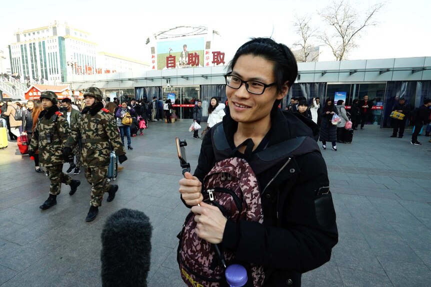 Xu Weifan holds a mobile phone and selfie stick while standing at the Beijing Train Station.