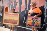 Two men in hats holding paintings in front of a mural with camels on it