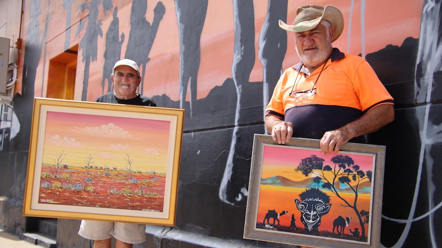 Two men in hats holding paintings in front of a mural with camels on it
