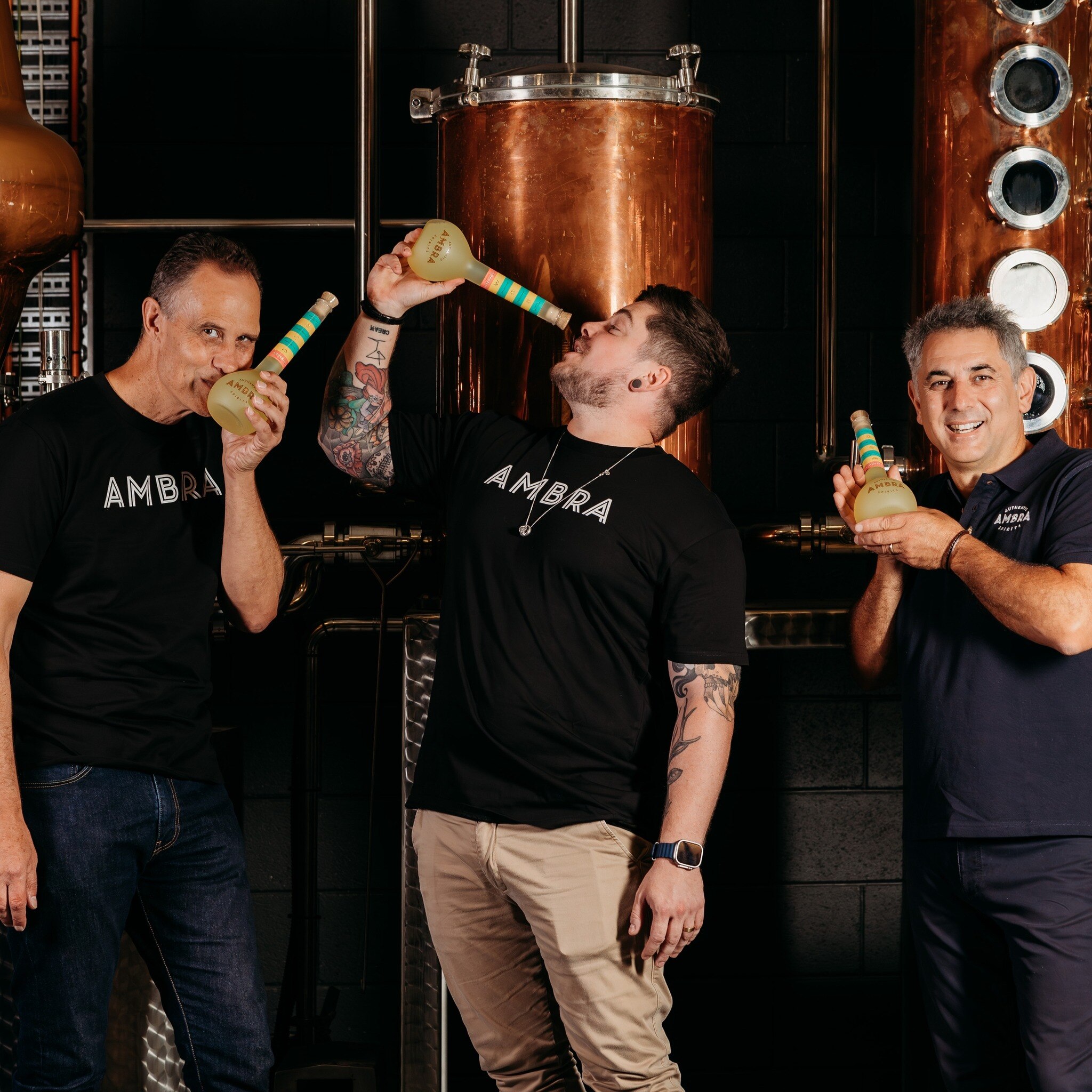 Tres hombres con camisas negras sostienen botellas de limoncello frente a alambiques de cobre. 