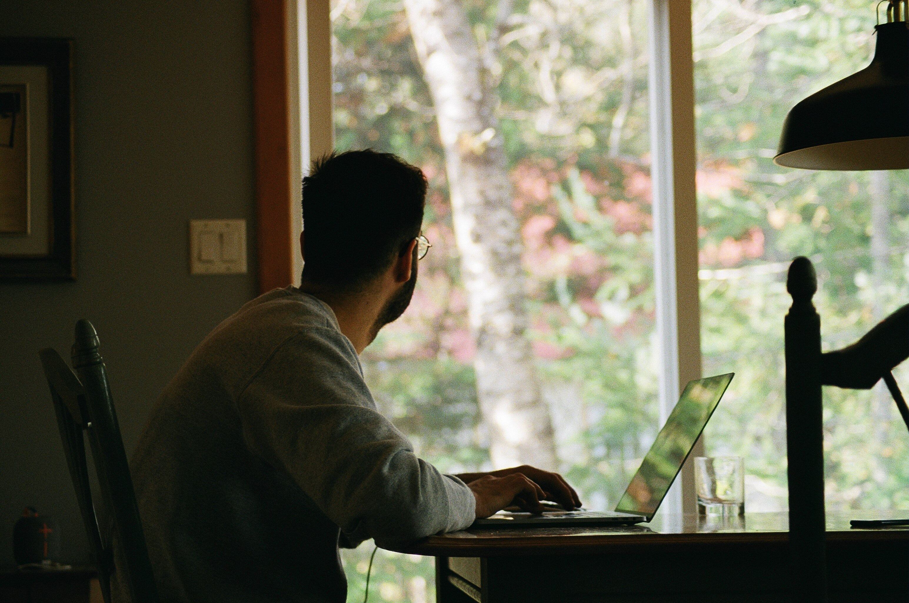 Man in darkened room sits at desk, typing at laptop and looking out window with back of head to camera.