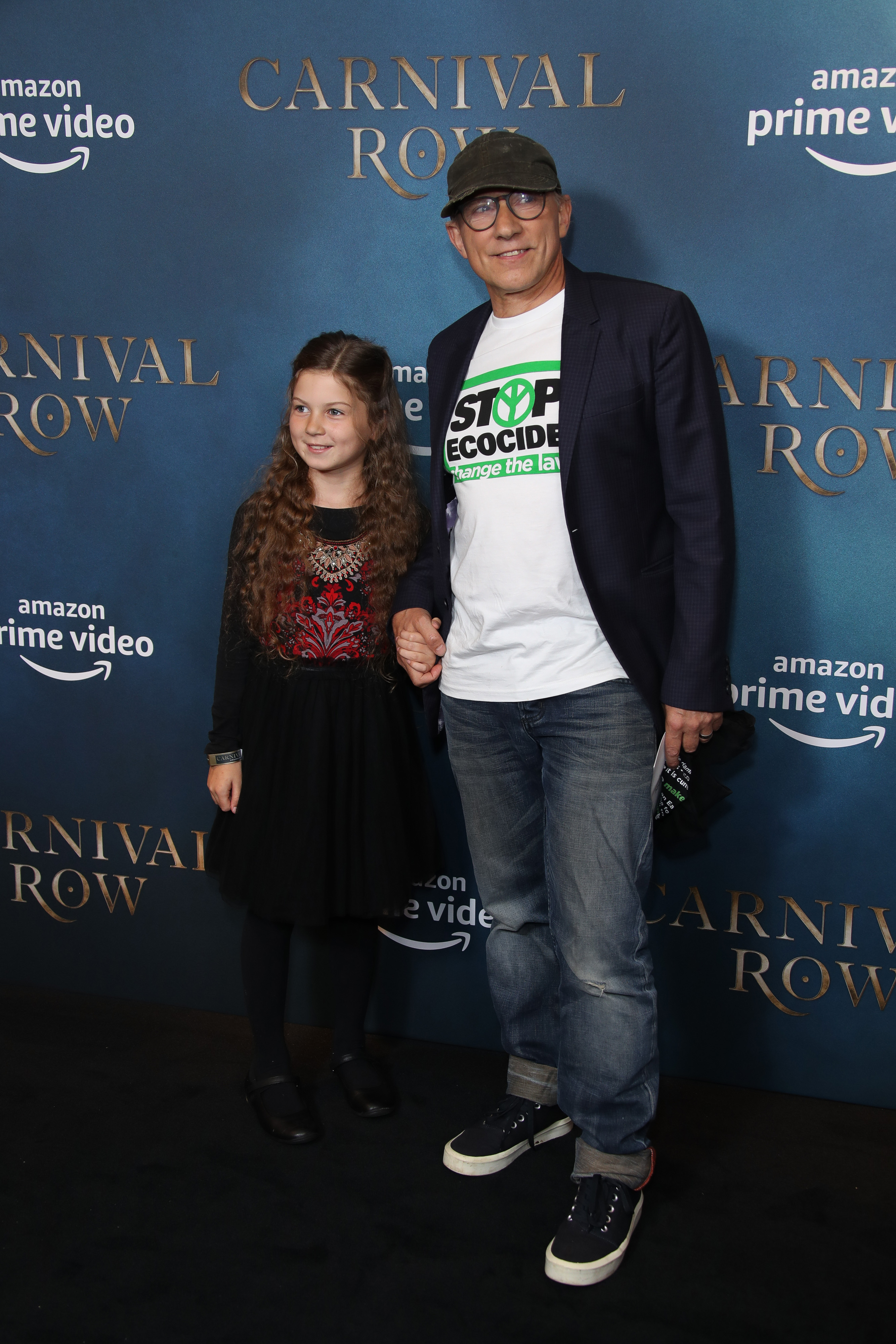 Simon McBurney, a middle-aged white man, wears a t-shirt reading "stop ecocide" on a red carpet with his young brunette daughter