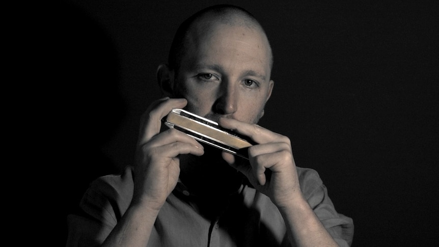 A high contrast photo of harmonica player Chris Maunders with his chromatic harmonica; he's looking into the camera