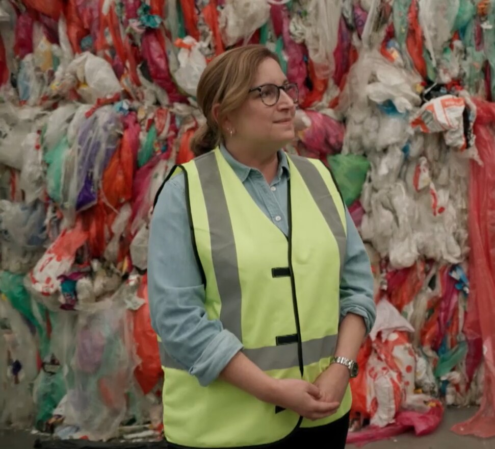 A woman with glasses wearing a high-vis vest stands in front of plastic waste piles.