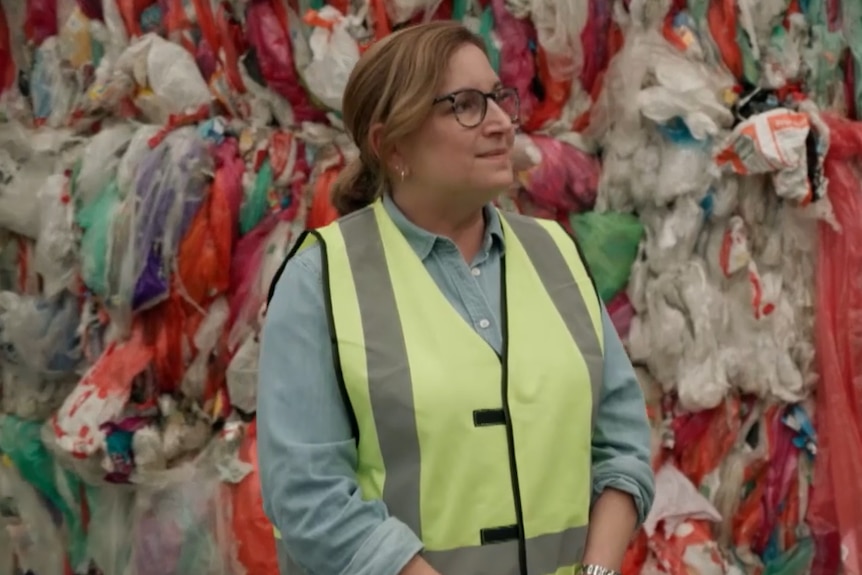 A woman with glasses wearing a high-vis vest stands in front of plastic waste piles.