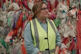 A woman with glasses wearing a high-vis vest stands in front of plastic waste piles.