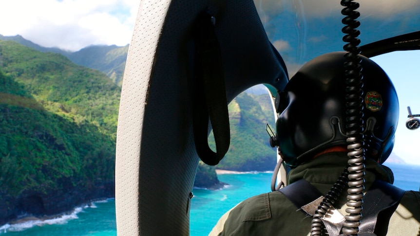 Looking from behind, you view a helicopter pilot as he looks over verdant green cliffs.