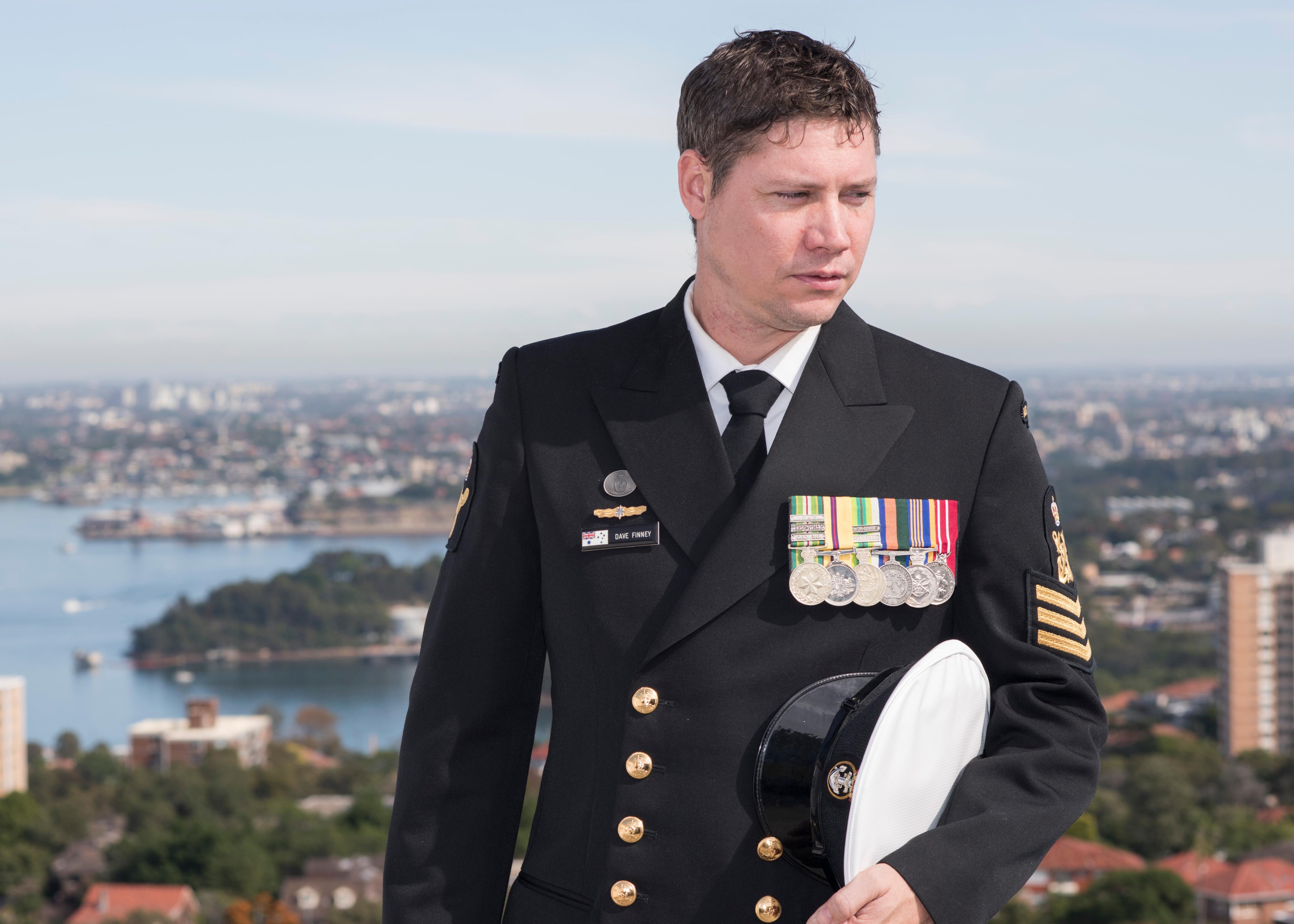 A young man in a military uniform wearing medals stands with his Navy cap under his arm.