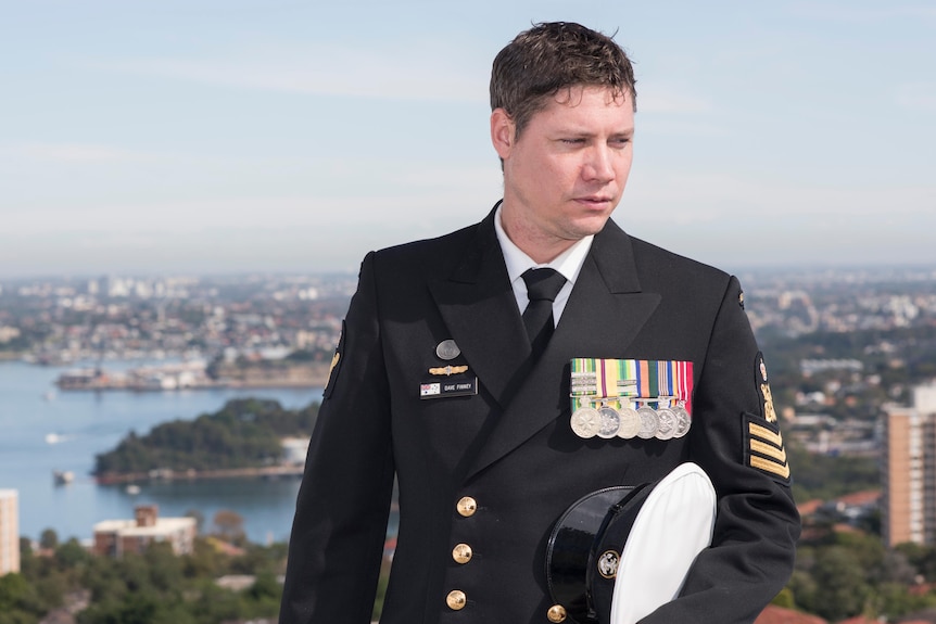 A young man in a military uniform wearing medals stands with his Navy cap under his arm.