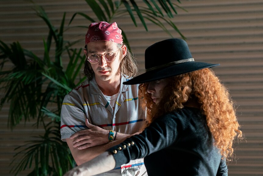 A man with round glasses and red bandana with arms crossed stands near woman with long red curly hair and black hat and jacket.