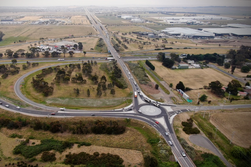 A drone image of traffic along a road with a roundabout going through fields, with development further away.