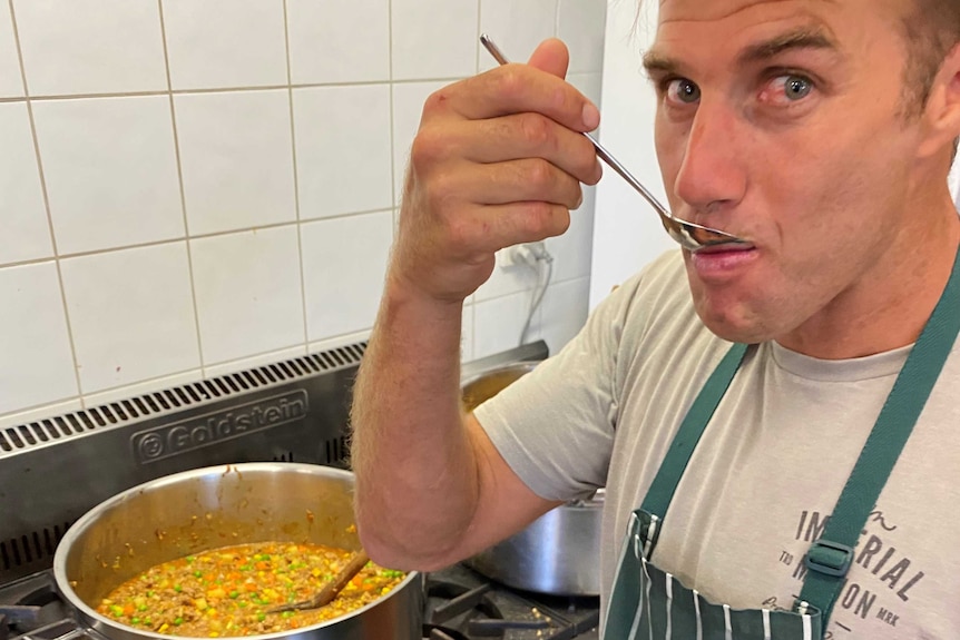 Man eating off spoon with pot of food behind him on stove.