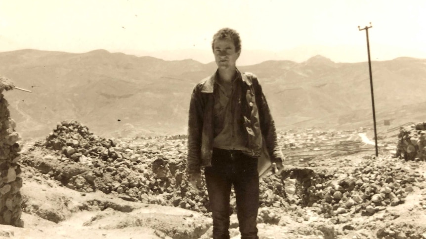 Black and white photo of a young man standing in front of a distant mountain ridge, with rough stone buildings in the foreground