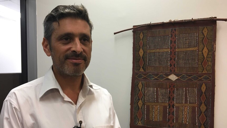 A man in a white coloured business shirt standing in an office in front of an Aboriginal artwork, smiling slightly.