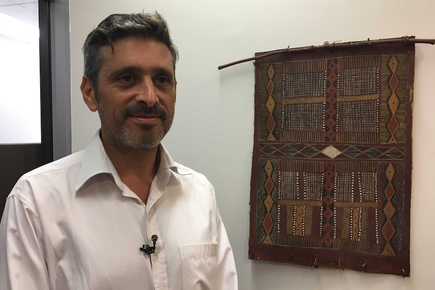 A man in a white coloured business shirt standing in an office in front of an Aboriginal artwork, smiling slightly.