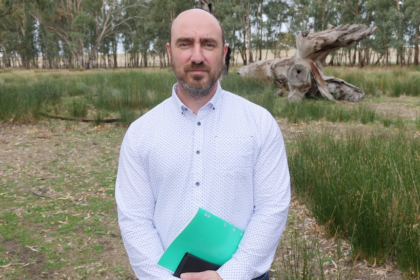 a man with a goatee and mustache wearing a white dotted shirt stands in a paddock in front of a log carrying a green folder