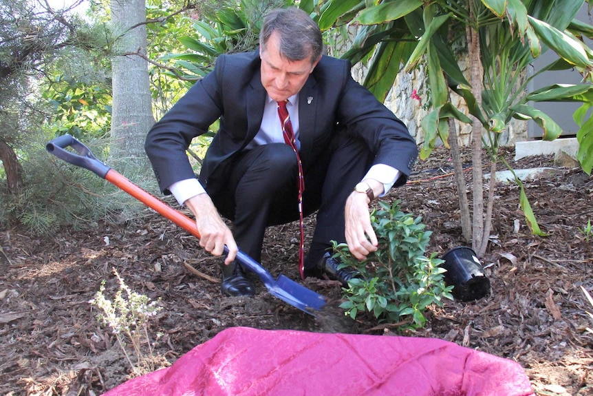 Graham Quirk, wearing a suit and tie, squats in a garden as he plants a tree behind the unveiled memorial.