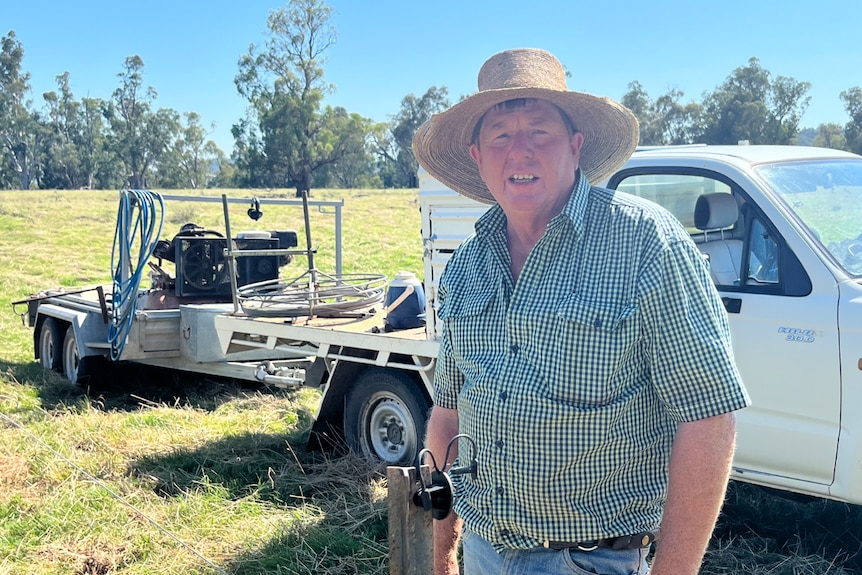 Man standing in a field, smiling at camera with ute and fencing equipment in background 